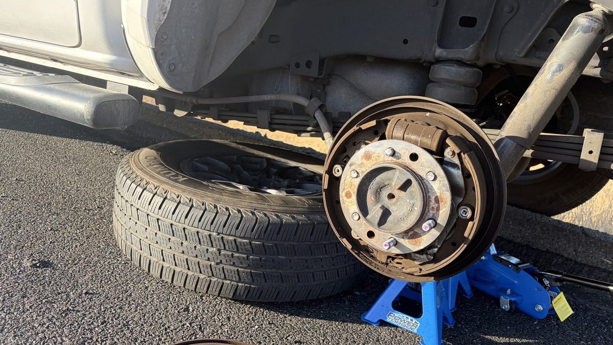 Rear brake drum pulled on a white pickup during a roadside brake job, with the wheel, jack stand and floor jack on the asphalt