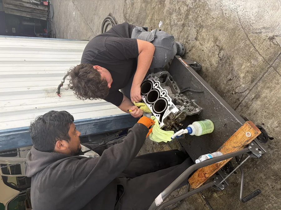 Two technicians cleaning a cylinder head on a bench outside