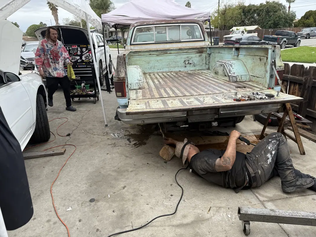 A mechanic lying under a pickup on a creeper in a driveway while another stands by the bed