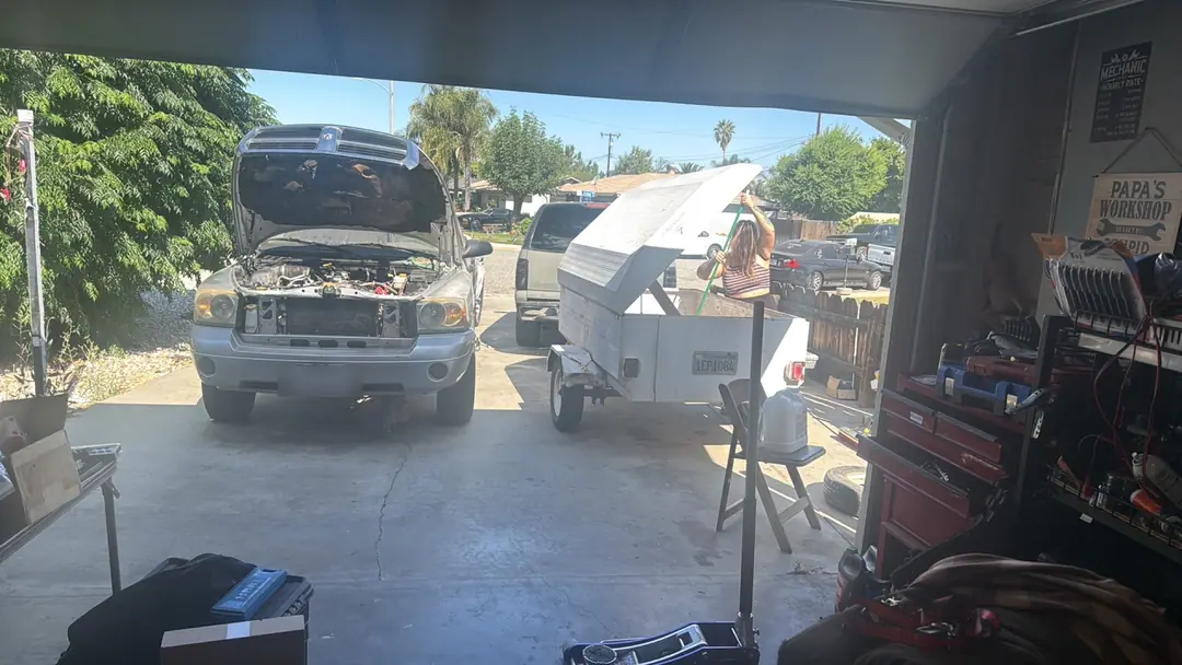A Chevy truck with its hood up in a garage while another vehicle waits outside, July 2025