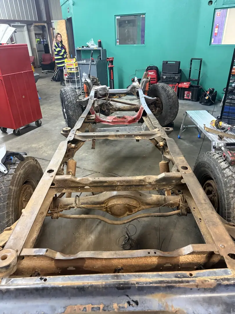 A bare pickup truck frame with axles and wheels inside a shop bay with green and purple walls