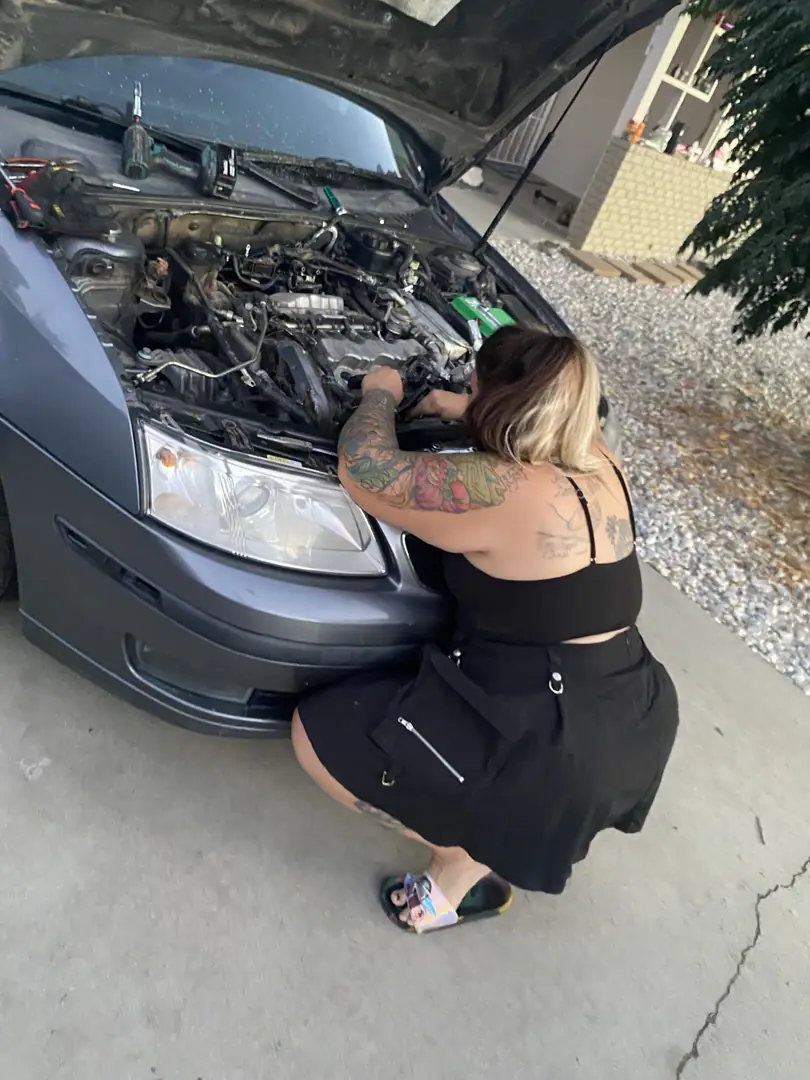 A mechanic crouched at the front of a gray Saab 9-3 with the hood open