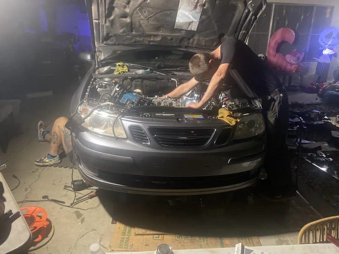 A gray Saab 9-3 with the hood up in a driveway at night, a mechanic leaning into the engine bay