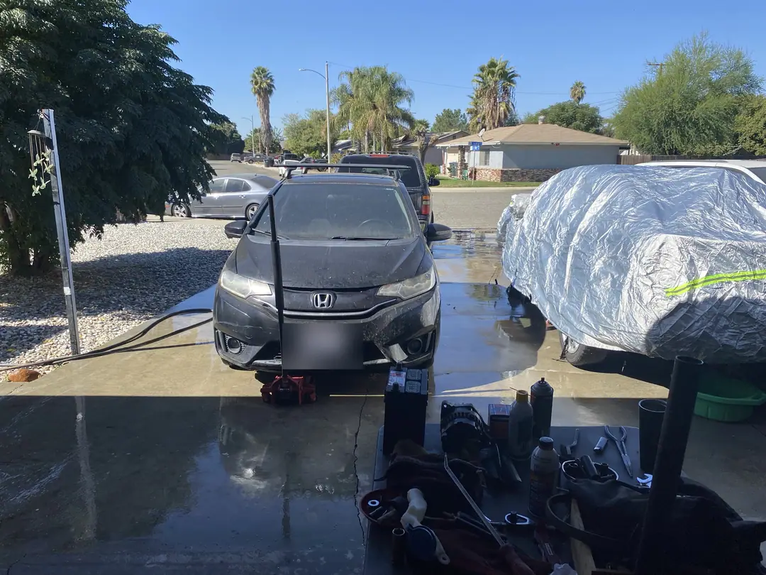 The driveway in November 2024 with a dark hatchback, a covered car and tools on the ground