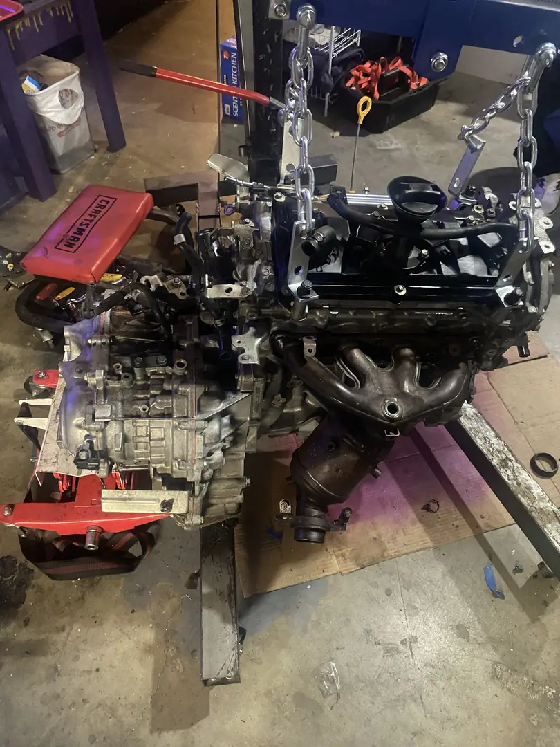 A four cylinder engine and transaxle hanging from a hoist in a garage