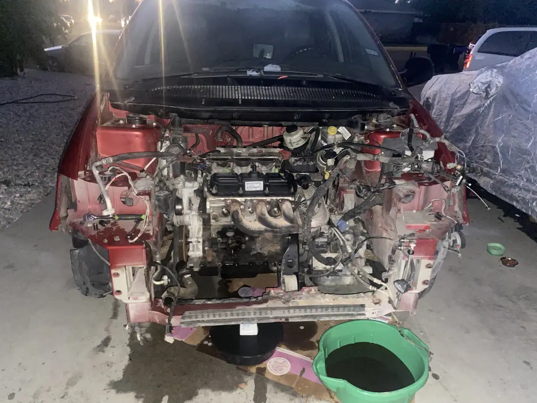 A red car in a driveway at night with the bumper and front end removed and the engine exposed, a bucket of drained coolant below