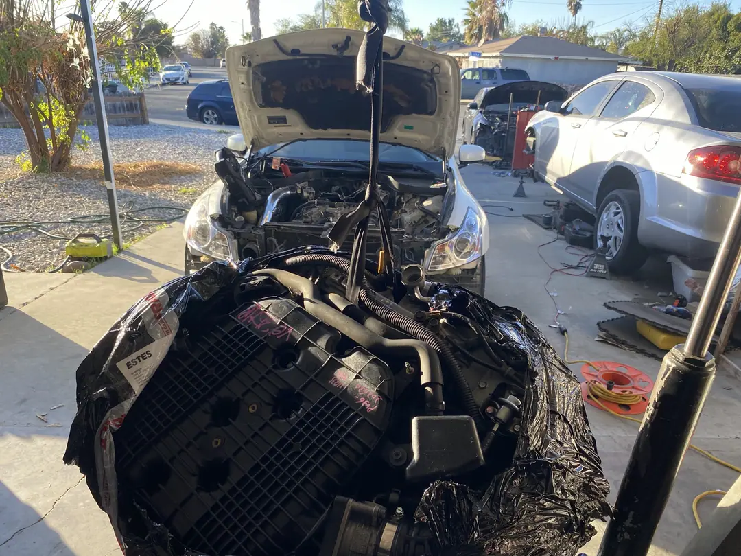 A used V6 engine wrapped in black plastic with a freight label, hanging from a hoist in front of a white sedan with the hood up