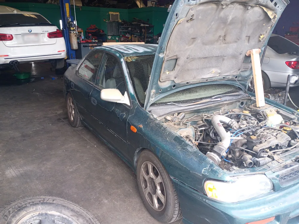 A green 1998 Subaru Impreza with the hood propped open inside a repair shop