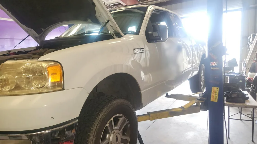 A white 2004 Ford F150 up on a two post lift with the hood open during a transmission swap