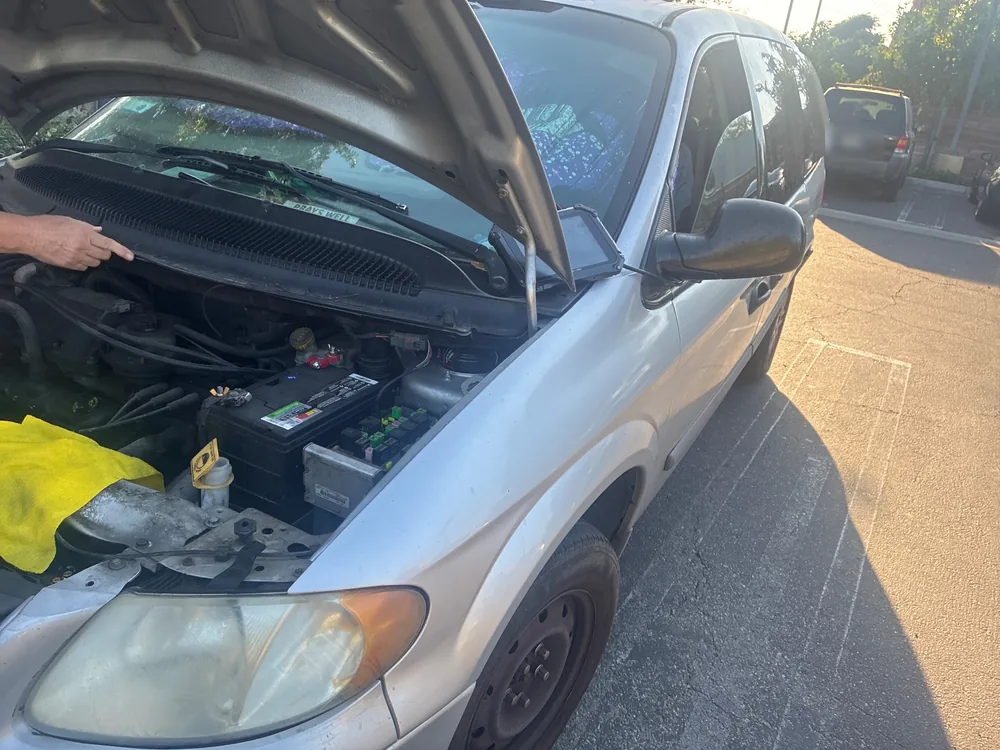 A silver 2006 Dodge Grand Caravan with the hood up in a parking lot while our tech points at the engine