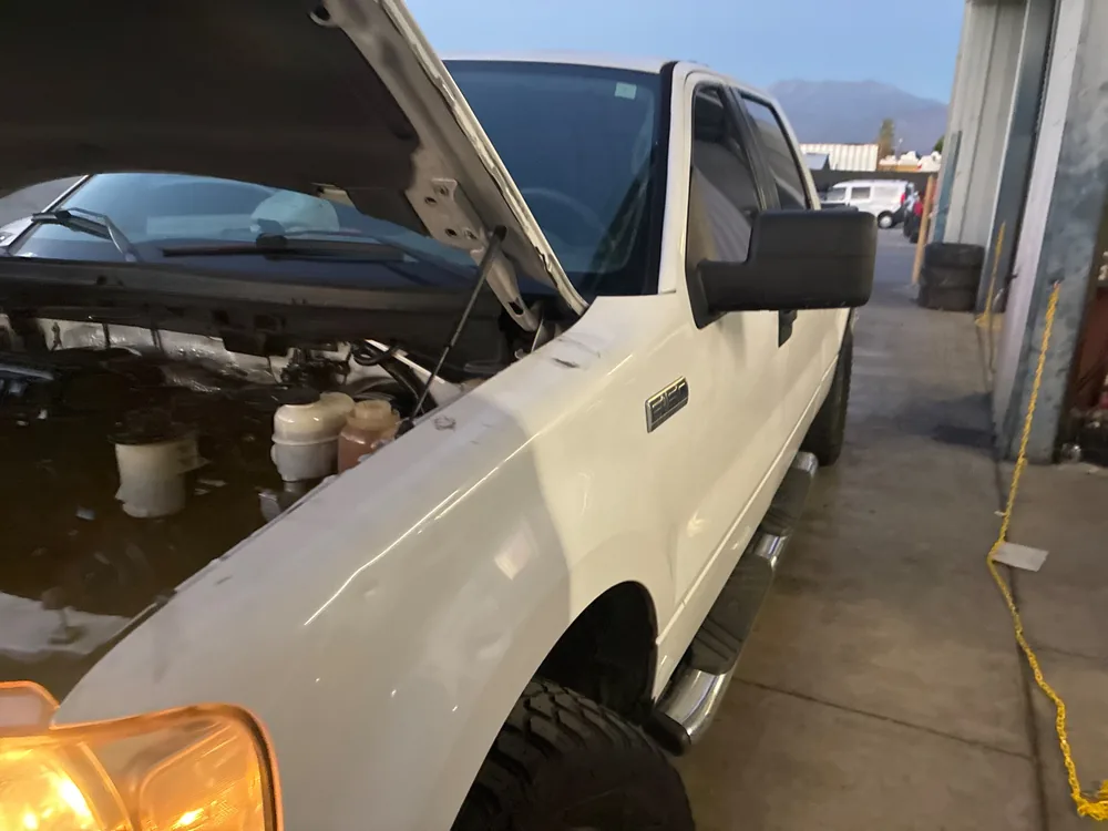 A white 2006 Ford F-150 with the hood up beside a shop bay